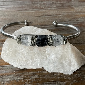 May include: A silver bangle bracelet with three stones set in a row. The stones are clear quartz, black onyx, and clear quartz. The bracelet is on a white background.