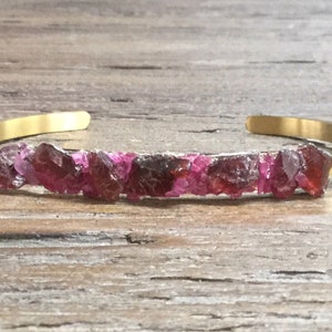 May include: A gold cuff bracelet with a row of pink and red gemstone chips.