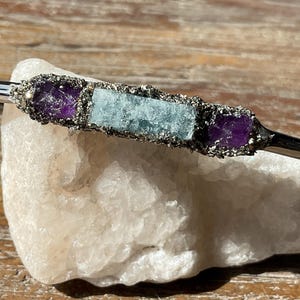 May include: A silver bangle bracelet with three stones set in silver. The stones are a light blue aquamarine and two purple amethysts.