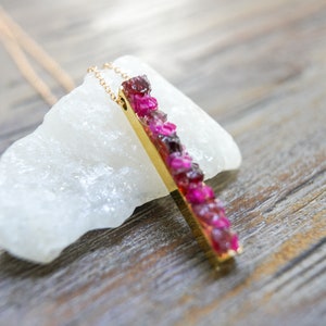 May include: A gold necklace with a pendant made of a long, thin bar of gold set with pink and red gemstones.