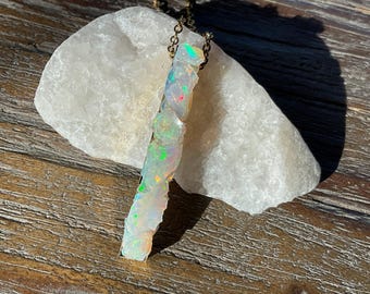 May include: A long, iridescent opal pendant hangs from a gold chain. The opal is a natural stone with a rough, textured surface. The pendant is a unique and eye-catching piece of jewelry.