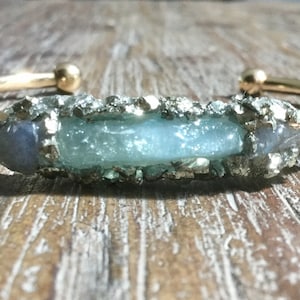 Op de afbeelding: Goudkleurige manchetarmband met een centrale, lichtblauwe edelsteen en een getextureerde, zilverkleurige rand. De armband heeft een eenvoudig, open ontwerp met afgeronde gouden accenten. De edelsteen is ongeveer 2.5 cm lang.