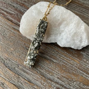 Puede incluir: Un collar de cadena dorada con un colgante rectangular largo hecho de pirita en bruto. El colgante tiene una superficie rugosa y texturizada y es de color gris oscuro.