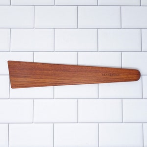 May include: A wooden spatula with a slightly curved shape and a brown wood grain. The spatula has a small, rectangular label that reads "RAINBOW WOOD".