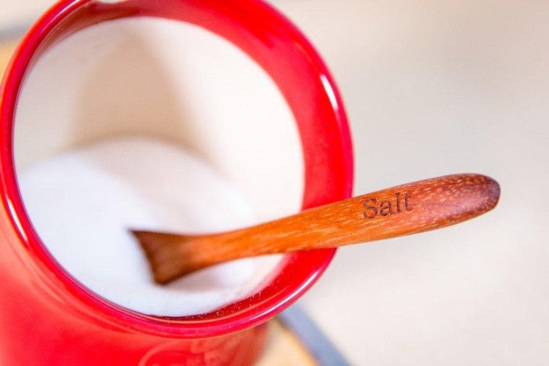 Puede incluir: Un cuenco de cer&aacute;mica rojo lleno de sal blanca y una cuchara de madera con la etiqueta "Sal" apoyada en el borde.