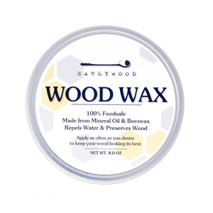 May include: A white round container with a silver lid. The label reads "EARLYWOOD WOOD WAX" and states that it is 100% foodsafe, made from mineral oil and beeswax, repels water and preserves wood. The label also states "Apply as often as you desire to keep your wood looking its best." and "NET WT. 8.0 OZ".