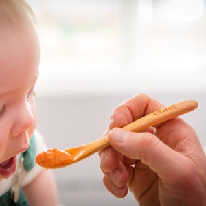 Puede incluir: Un beb&eacute; est&aacute; siendo alimentado con pur&eacute; de naranja de una cuchara de madera con la palabra "Clark" grabada en ella.
