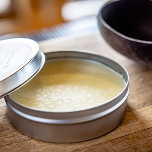 May include: A silver tin with a lid, containing a pale yellow wax. The lid has the word "WOODWAX" printed on it.