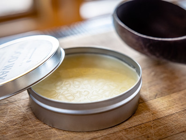 May include: A silver tin with a lid, containing a pale yellow wax. The lid has the word "WOODWAX" printed on it.