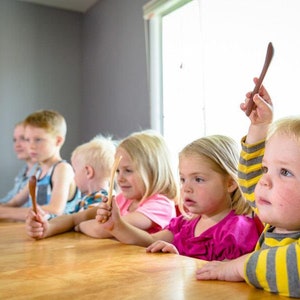 Puede incluir: Cinco ni&ntilde;os est&aacute;n sentados en una mesa de madera, cada uno sosteniendo una cuchara de madera. El ni&ntilde;o en primer plano levanta su cuchara en el aire.
