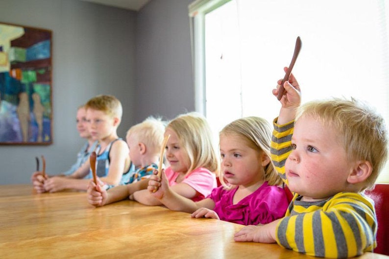 Puede incluir: Cinco ni&ntilde;os est&aacute;n sentados en una mesa de madera, cada uno sosteniendo una cuchara de madera. El ni&ntilde;o en primer plano levanta su cuchara en el aire.