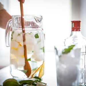 May include: A glass pitcher filled with ice cubes, water, and fresh mint leaves. A wooden spoon is stirring the mixture. A clear glass bottle with a red lid is in the background.