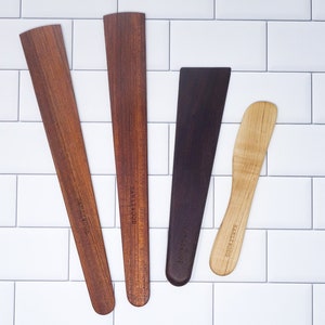 May include: Four wooden kitchen utensils with the text "Sallywood" engraved on each. The utensils are a brown spatula, a brown butter knife, a dark brown spatula, and a light brown butter knife.