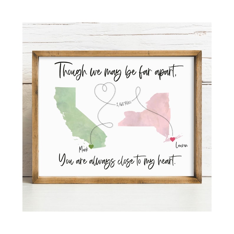 K&ouml;nnte beinhalten: Ein gerahmter Druck mit einer gr&uuml;nen Aquarellkarte von Kalifornien und einer rosa Aquarellkarte von New York. Eine gepunktete Linie verbindet beide in Herzform mit dem Text 2,468 Miles. Der Text lautet Though we may be far apart, You are always close to my heart, mit den Namen Mark und Lauren.