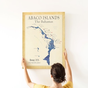 May include: A framed map of Abaco Islands, The Bahamas, with a blue watercolour design. The map includes the text "Abaco 2025 Our favourite place" and is held by a person.