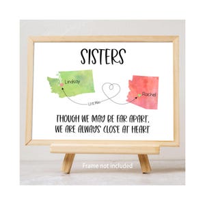 Può includere: Una stampa incorniciata su un cavalletto di legno. La stampa presenta la parola "SISTERS" sopra due mappe statali ad acquerello, una verde e una rossa, collegate da una linea a forma di cuore. Il testo recita: "THOUGH WE MAY BE FAR APART, WE ARE ALWAYS CLOSE AT HEART."