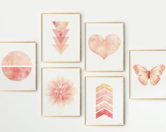 May include: A set of six framed wall art prints featuring watercolour designs in shades of pink and peach. The prints display a circle, stacked triangles, a heart, a flower, stacked chevrons, and a butterfly. Each piece is in a thin gold frame.
