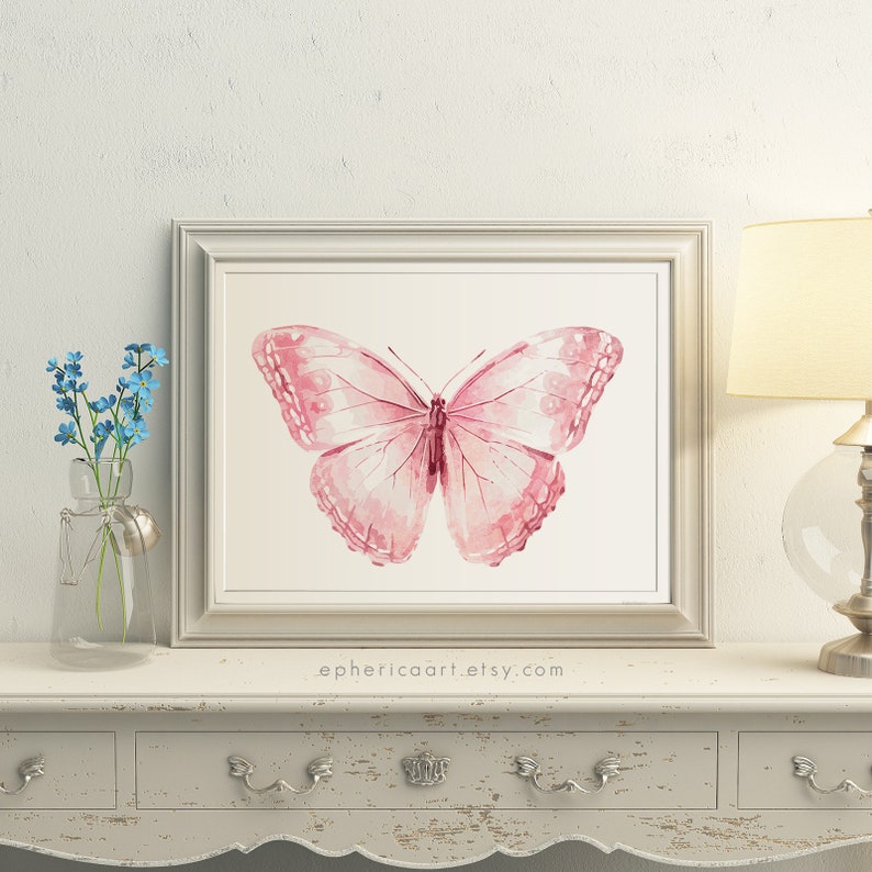 May include: A framed watercolor print of a pink butterfly. The butterfly has detailed wing patterns and is centered on a cream-colored background. The frame is a light silver color. The text "ephericaart.etsy.com" is at the bottom.