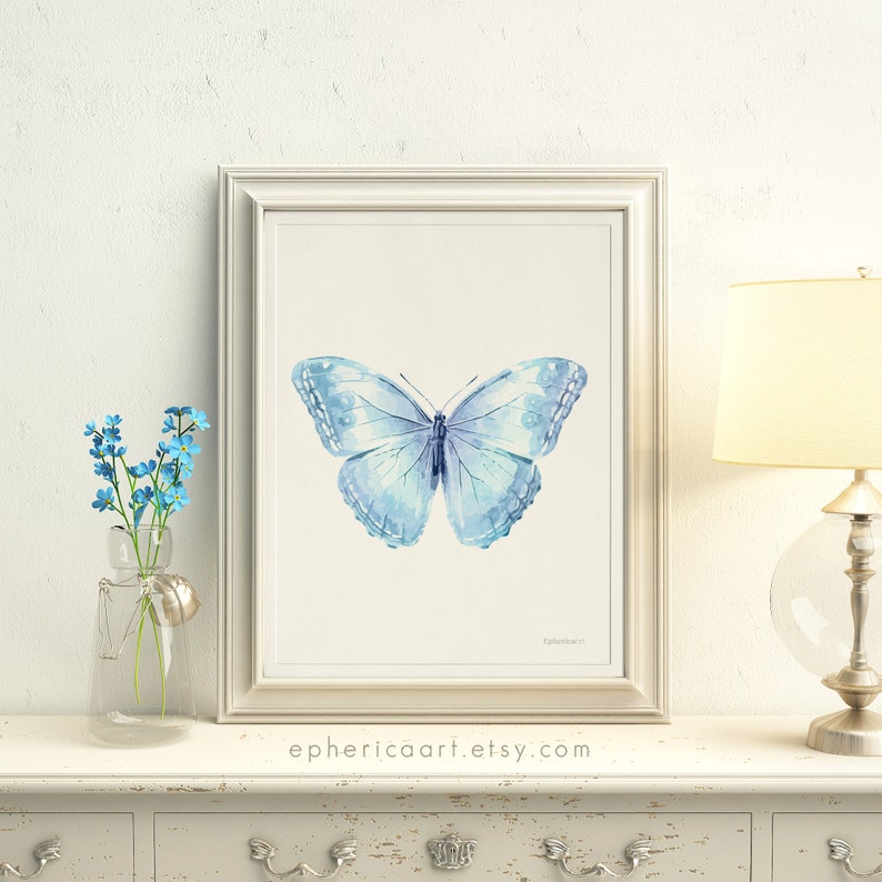 May include: A framed watercolor print of a light blue butterfly with delicate wing details. The artwork is displayed on a white dresser, next to a vase of blue flowers. The website address ephericaart.etsy.com is printed at the bottom.