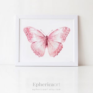 May include: A framed watercolor print featuring a pink butterfly with detailed wing patterns. The butterfly is centered on a white background within a white frame. The print has a soft, artistic aesthetic. The artist's name and website are at the bottom.
