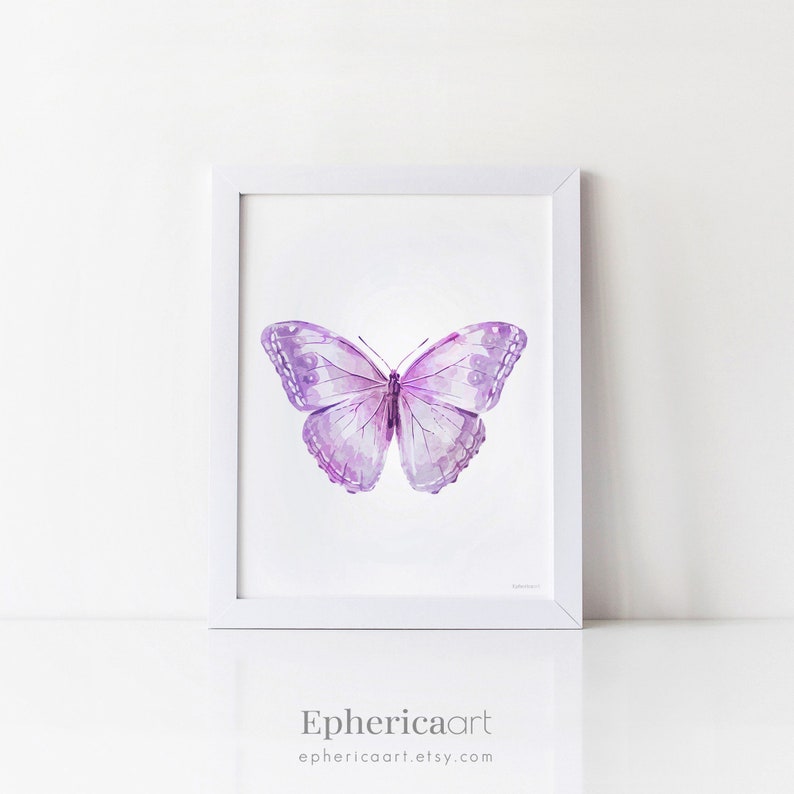 May include: A framed watercolor print of a butterfly. The butterfly is depicted with shades of purple and lavender, with detailed wing patterns. The artwork is set against a white background within a white frame. The artist's name and Etsy shop URL are at the bottom.