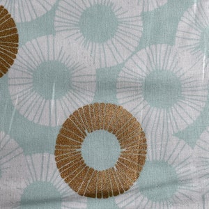 May include: A white fabric with a light blue and gold pattern. The pattern features a repeating design of stylized sunbursts with a gold ring in the center.