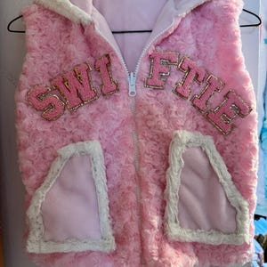May include: Pink faux fur vest with a zipper closure and a white trim. The vest has two pockets and the word "SWIFTIE" is embroidered on the front in pink and gold letters.