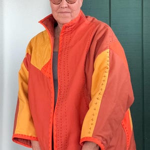 May include: A quilted jacket in shades of orange and yellow. The jacket features a stand-up collar, a zipper closure, and contrasting stitching details. The sleeves are wide and the jacket appears to be handmade.