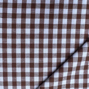 May include: A fabric featuring a classic gingham pattern with a repeating grid of small, solid brown squares alternating with white squares. The material is folded over at the bottom right corner, revealing the texture of the woven textile surface.