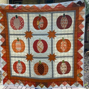 May include: A white quilt with a brown border and a patchwork design featuring orange, brown, and white pumpkins. The pumpkins are arranged in a grid pattern and have different patterns.