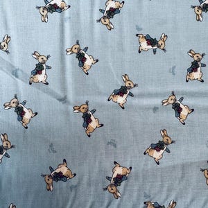 May include: Light blue fabric with a repeating pattern of cartoon rabbits wearing jackets. The rabbits are in various poses, with brown and white fur, and the jackets are blue and green. Small floral accents are scattered throughout.