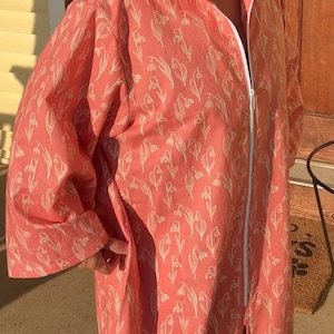 May include: A coral-colored robe with a white floral pattern and a white zipper. The robe has long sleeves and a collared neckline. The fabric appears to be lightweight, suitable for warmer weather. The robe is knee-length.