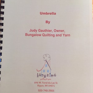 May include: The cover of a quilting pattern book titled "Umbrella" by Judy Gauthier, Owner, Bungalow Quilting and Yarn. The book cover features a stylized illustration of a house with a chimney and the text "bungalow quilting & yarn". The address, phone number, and website for Bungalow Quilting and Yarn are also included on the cover.