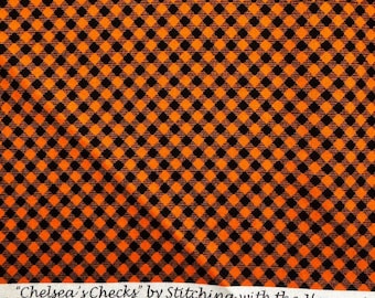 May include: Orange and black checkered fabric. The fabric has a repeating pattern of orange and black squares. The text "Chelsea's Checks" by Stitching with the Housewife is printed on a white strip of fabric. A purple ruler is at the bottom.