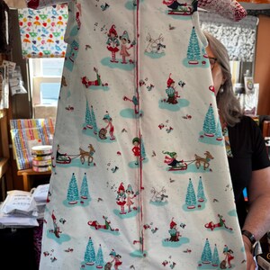 May include: A white baby sleep sack with a festive Christmas-themed pattern. The design features elves, reindeer, Christmas trees, and musical notes in red, green, and blue. The sleep sack has a full-length zipper and is displayed on a hanger.