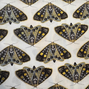 May include: White fabric with a repeating pattern of stylized moths. Each moth features black, gold, and silver details, with a symmetrical design. The moths have a detailed wing pattern and are evenly spaced across the fabric.