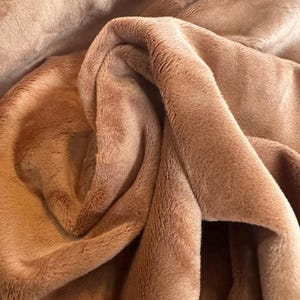 May include: Close-up of a soft, plush fabric in a warm, light brown color. The material is folded and draped, showing the texture and depth of the fabric. The fabric appears to be faux fur or a similar material.