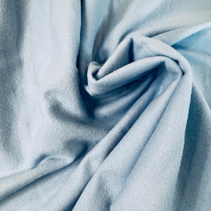 May include: A close-up shot of a light blue fabric with a soft, textured appearance. The fabric is gathered and folded, creating shadows and highlights that emphasize its texture. The color is a pale, muted blue.
