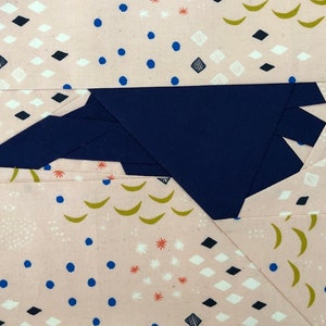 May include: A close-up of a fabric patchwork design. The design features a large, dark blue geometric shape set against a light pink background. The background is decorated with small blue dots, black diamonds, and gold crescent shapes.