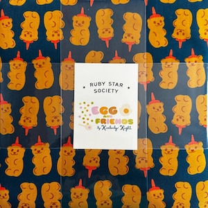 May include: A fabric square featuring a repeating pattern of orange gummy bear illustrations on a dark blue background. Each bear has a red hat and is on a red stick. A white label reads "RUBY STAR SOCIETY" and "EGG AND FRIENDS by Kimberly Knight."