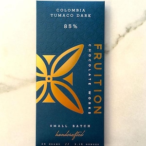 May include: A dark blue chocolate bar wrapper with gold lettering and a gold design. The wrapper says "COLOMBIA TUMACO DARK 85% FRUITION CHOCOLATE WORKS SMALL BATCH handcrafted 60 GRAMS // 2.12 OUNCES".