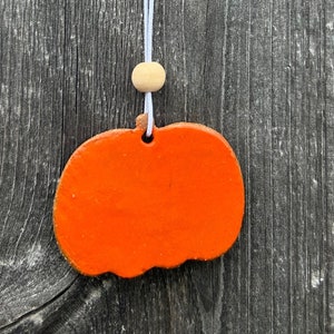 May include: A bright orange, flat, pumpkin-shaped ornament hanging from a thin white string. A small, round, light-colored wooden bead is threaded onto the string above the pumpkin. The pumpkin has a smooth, solid-colored surface and a hole at the top.