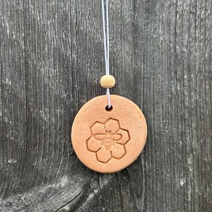 May include: A circular terracotta-colored clay ornament featuring an embossed bee and honeycomb design. The ornament is suspended by a thin white cord with a single light-colored wooden bead above the clay disc, hanging from a metal screw.