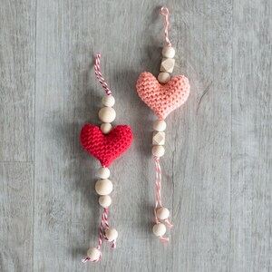 Könnte beinhalten: Zwei gehäkelte Herz-Anhänger mit Holzperlen und roter und weißer Schnur. Die Herzen sind rosa und rot.
