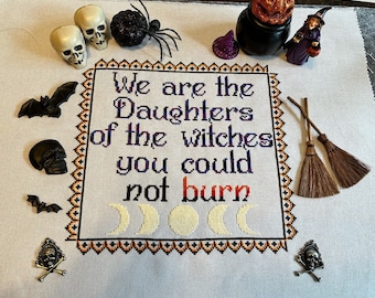 May include: Cross-stitch embroidery with a black and white border and a white background. The text reads "We are the Daughters of the witches you could not burn". There are three yellow moons in the center of the design.
