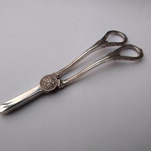 May include: Silver serving tongs with a decorative floral motif on the central joint. The tongs feature looped handles and a gently curved design. The overall aesthetic is vintage and elegant.