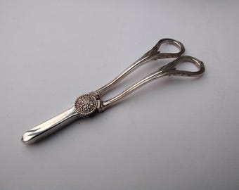 May include: Silver-toned serving tongs with a decorative floral design on the central joint. The tongs have looped handles and a slightly curved design. The overall aesthetic is vintage and elegant.
