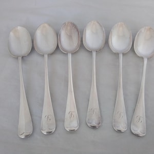 May include: Six silver spoons with oval bowls and long, tapered handles. Each spoon has a monogrammed initial "F" engraved on the handle. The spoons are arranged in a row on a light gray surface.