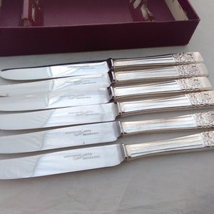 Puede incluir: Juego de seis cuchillos de mantequilla de plata con mangos ornamentados. Los cuchillos est&aacute;n en su caja original. La caja es de color burdeos con letras blancas. Los cuchillos est&aacute;n estampados con "SUPER STAINLESS QUALITY" y "CO-OP" con "SWITZERLAND MADE".