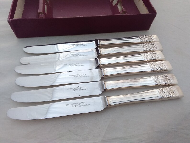 Puede incluir: Juego de seis cuchillos de mantequilla de plata con mangos ornamentados. Los cuchillos est&aacute;n en su caja original. La caja es de color burdeos con letras blancas. Los cuchillos est&aacute;n estampados con "SUPER STAINLESS QUALITY" y "CO-OP" con "SWITZERLAND MADE".